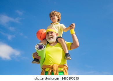 Happy Grandfather Giving Grandson Piggyback Ride On His Shoulders And Looking Up. Grandfather And Grandson With Basketball Ball And Yoga Mat In Hands