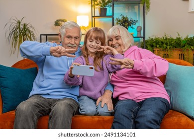 Happy grandfather, grandmother and granddaughter taking selfie photo with smartphone at home. Smiling Caucasian girl with grandparents make blogger self portrait showing peace sign sitting on sofa. - Powered by Shutterstock