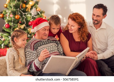 Happy grandfather is showing photos in album to his children. They are sitting on couch near Christmas tree and smiling Stock Photo