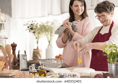 Happy Grandma Cracking An Egg And Cooking In A Kitchen With Her Granddaughter