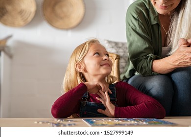 Happy Grandmother And Beautiful Granddaughter Looking At Each Other While Solving Puzzle. Smiling Girl Trying To Solve Jigsaw Puzzle Asking Help To Old Granny. Child Playing At Home With Senior Woman.