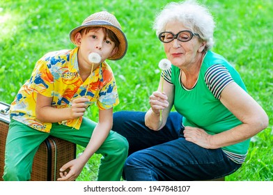 Happy Grandmother And Grandchild Hold Dandelions Pretending To Be Singing Into A Microphone. Older Woman And Boy Fooling Around While On A Green Lawn On Suitcases. Family Relationships. Fun In Spring