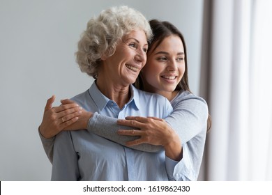 Happy Grandmother And Granddaughter Hugging, Standing At Home, Looking Out Window, Beautiful Girl Grandchild Expressing Love And Care, Embracing Grandma, Two Generations, Good Relationship