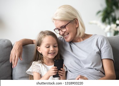 Happy grandmother and granddaughter using phone together, sitting on cozy sofa at home, browsing mobile device apps, grandma with grandchild playing game, looking at screen, having fun