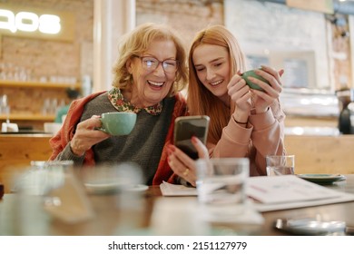 Happy grandmother and her adolescent granddaughter sitting in a cafe, enjoying coffee and using smart phone for funny videos. - Powered by Shutterstock