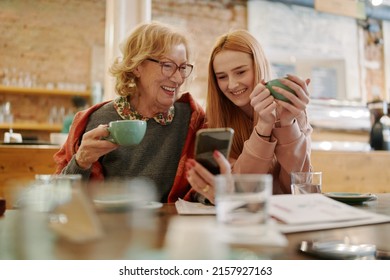 Happy grandmother and her adolescent granddaughter sitting in a cafe, enjoying coffee and using smart phone for funny videos. - Powered by Shutterstock