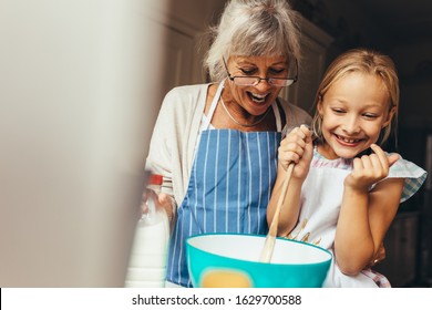 Happy Grandmother And Kid Mixing A Cake Batter At Home. Happy Little Girl Stirring Batter In A Bowl With Her Granny Standing By Her Side.