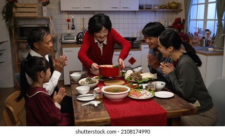 Happy Grandmother Serving Delicious Food On Dining Table As Her Family Members Clapping Hands For Her. Getting Ready For Big Meal On Chinese New Year's Eve