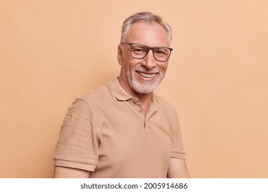 Happy grandpa senior man with grey hair and beard smiles toothily shows perfect white teeth wears optical glasses and casual t shirt enjoys life glad to meed old friend isolated over beige background