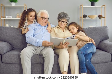 Happy grandparents and their two granddaughters look at a photo book with a family photo together. Grandparents and little girls at home sitting on the couch in the living room. Family concept. - Powered by Shutterstock