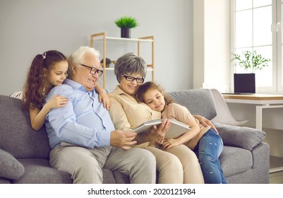 Happy Grandparents With Twin Grandchildren Browse Family Photo Album And Share Happy Memories. Family Having Fun At Home Sitting On Sofa In Living Room. Concept Of Family History And Memories.