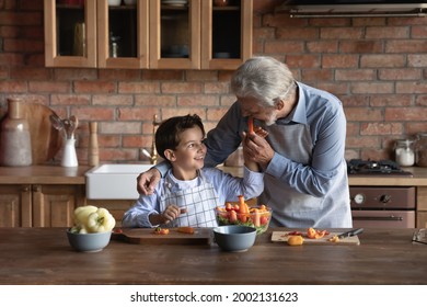 Happy grandson and grandfather engaged in cooking salad for lunch at home, cutting fresh vegetables at kitchen table, keeping healthy diet,, tasting slice of pepper. Family eating concept - Powered by Shutterstock