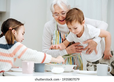 Happy granny holding her grandson from falling. He is standing on a stool, reaching for the food. His sister also grabing herself some.