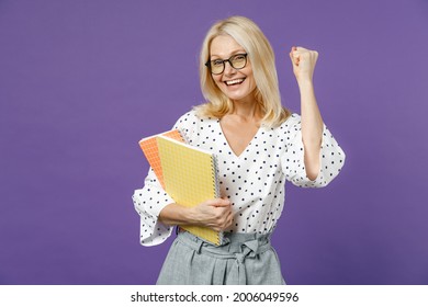 Happy Gray-haired Blonde Teacher Woman Lady 40s 50s Years Old In White Dotted Blouse Eyeglasses Standing Hold Notepads Doing Winner Gesture Isolated On Bright Violet Color Background Studio Portrait