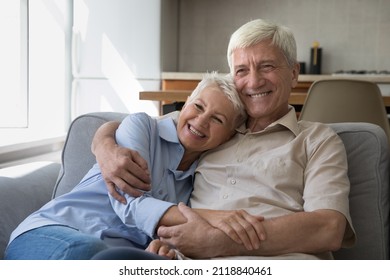 Happy Grey Haired Mature Couple Home Candid Portrait. Positive Smiling Senior Husband And Wife Resting On Couch At Home, Hugging With Love, Care, Tenderness, Looking Away, Smiling, Laughing