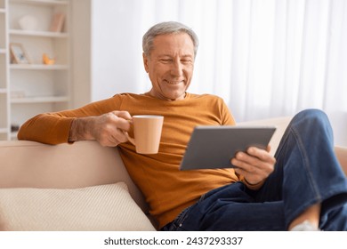 Happy grey-haired elderly man sitting on couch at home, drinking coffee and using digital tablet, watching movie or favorite podcast, grandfather enjoying digital world - Powered by Shutterstock