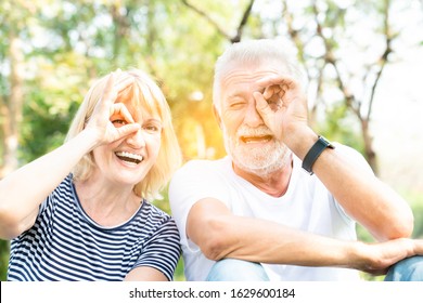 Happy Grey-haired Old Man Wearing T-shirt And Senior Woman Wearing Striped T-shirt Make Selfie In The Park With Happy Face Smiling Doing Ok Sign With Hand On Eye Looking Through Fingers.Body Language.