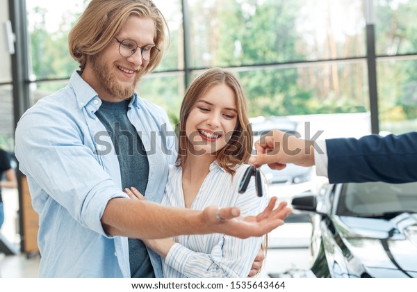 Happy and grinning young\
adult man hugging cheerful woman, looking at salesman, standing in\
auto salon. Confident dealer giving key from new rental car to\
smiling couple
