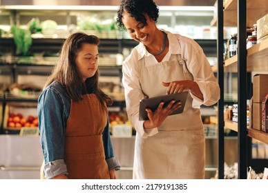 Happy Grocery Store Manager Using A Digital Tablet While Having A Discussion With Her Employee. Female Shop Owner Giving A Woman With Down Syndrome Training In Managing Online Grocery Orders.