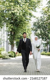 happy groom holding hands with smiling muslim bride with wedding bouquet and walking together