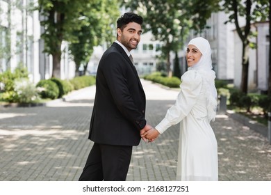 happy groom holding hands with smiling muslim bride in wedding dress and walking together