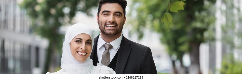 happy groom hugging muslim bride in hijab smiling and looking at camera, banner
