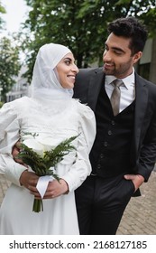 happy groom posing with hand in pocket and looking at muslim bride in hijab with wedding bouquet