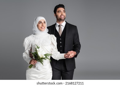 happy groom in suit holding hand of dreamy muslim bride with wedding bouquet of calla lily flowers isolated on grey