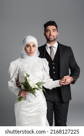 happy groom in suit holding hand of muslim bride with wedding bouquet of calla lily flowers isolated on grey