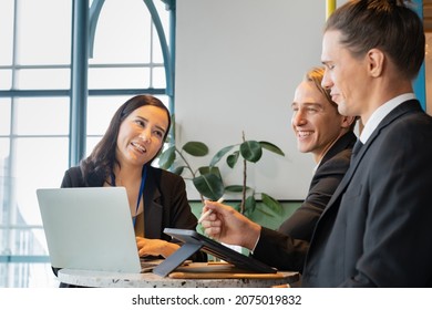 A Happy Group Of Business People, Asian Businesswoman And Caucasian Businessmen Sitting At A Round Table With Laptop And Tablet, Having A Business Conversation During The Small Conference In A Cafe.