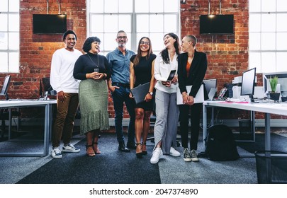 Happy group of businesspeople laughing cheerfully in a modern workplace. Diverse group of colleagues enjoying working together in an office. Successful businesspeople standing together. - Shutterstock ID 2047374890