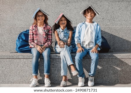 Happy group of elementary middle school kids children pupils classmates with backpacks holding notebooks on heads and having fun outdoors sitting on the stairs steps in school background