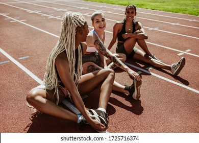 Happy Group Of Female Athletes Sitting On A Running Track And Stretching Legs. Women Runners Doing Warmup Exercises At The Stadium.