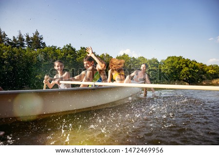 Happy group of friends having fun while laughting and swimming in river. Joyful men and women in swimsuit in a boat at riverside in sunny day. Summertime, friendship, resort, weekend concept.