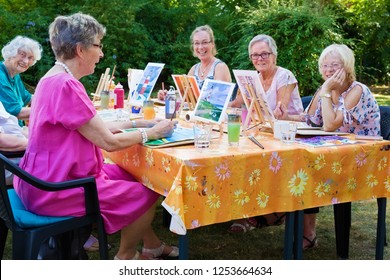 Happy Group Of Senior Ladies Enjoying Art Class Seated Around A Table Outdoors In The Garden Painting With Water Colors While Smiling And Chatting.