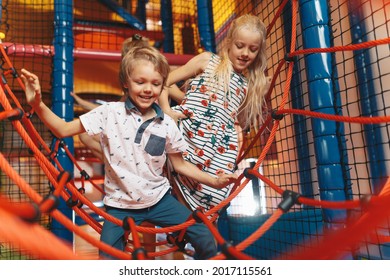 Happy group of siblings playing together on indoor playground. Excited kids playing together on net ropes. Cute school kids playing on the colorful playground at shopping mall