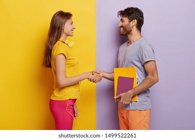 Happy groupmates meet after holidays, shake hands, agree to work together as team, stand in profile, lovely woman with headphones meets friend. Unshaven young man holds notepad talks with female