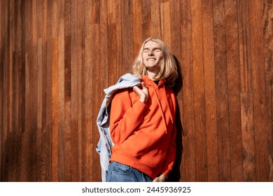 Happy guy basks in warm rays of sun while walking outdoor. Cheerful long-haired blond man leaning effortlessly against the wooden wall with eyes closed in bliss savouring every moment of serenity joy.