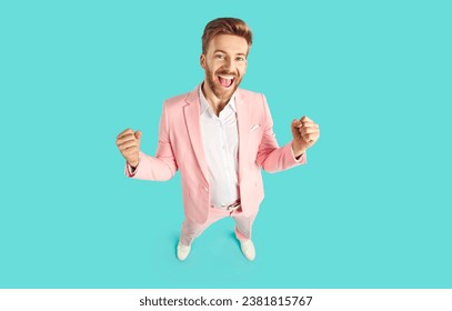 Happy guy celebrating his success and having fun. High angle shot of joyful young man in pink suit standing on blue color background, looking at camera, smiling and doing YES gesture. Fashion concept