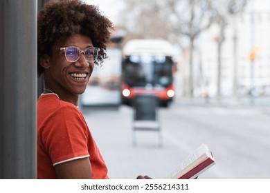 Happy guy with curly hair in casual clothes and eyeglasses reading book while waiting for bus on city street - Powered by Shutterstock