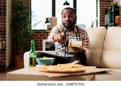 Happy Guy Holding Noodles Delivery Box And Switching Tv Channels With Remote Control To Find Movie. Eating Fast Food Takeout Meal With Chopsticks And Watching Film On Television, Having Fun.