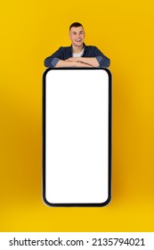 Happy Guy Leaning On Huge Smartphone With Empty Screen Advertising Great Mobile Application Smiling To Camera Over Yellow Background, Studio Shot. App Advertisement Concept. Vertical, Mockup