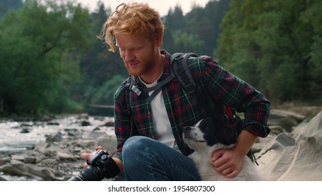 Happy Guy Petting Dog Outdoor. Smiling Hiker Enjoying Natural Landscape. Redhead Man With Dog Sitting At Mountain River. Male Photographer Giving Dog To Sniff Photo Camera