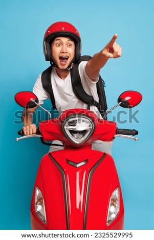 Happy guy in red helmet posing on red scooter, one hand on handle bar, another points ahead, fast drive concept, copy space