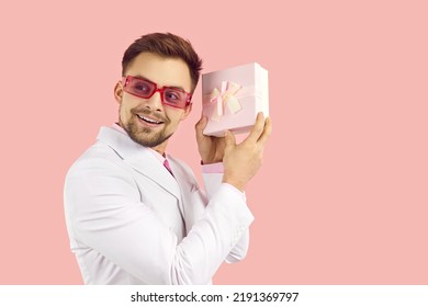 Happy Guy Shaking Present Trying To Guess What's Inside. Man In White Suit And Pink Sunglasses Standing Isolated On Pastel Pink Background, Holding Gift Box And Listening To Sound Coming From Inside