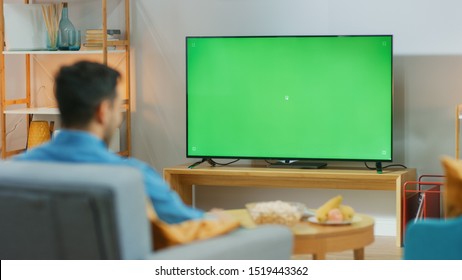 Happy Guy Sitting At Home In The Living Room On His Chair, Watching Green Chroma Key Screen, Relaxing After Work. Man In A Cozy Room Watching Sports Match, News, TV Show Or Movie.