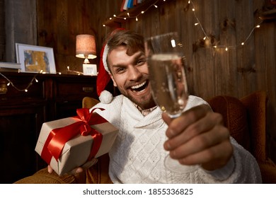 Happy Guy Wearing Santa Hat Holding Christmas Gift And Champagne Glass Saying Cheers Looking At Camera In Virtual Chat With Friends Or Family Celebrating New Year Distance Online Party. Web Cam View