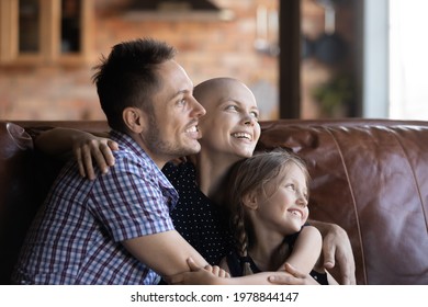 Happy Hairless Young Woman With Cancer Relaxing On Couch With Family, Hugging Husband And Little Daughter, Looking Away With Hope, Smiling, Thinking Of Good Optimistic Future. Oncology Concept