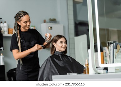 happy hairstylist spraying hair of female client, hairdresser with braids holding spray bottle near woman with short brunette hair in salon, haircut, hair treatment, hair make over, hairdo - Powered by Shutterstock