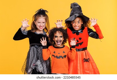 Happy Halloween! Cheerful Kids In Carnival Costumes And Makeup Make A Terrible Gesture On Bright Colored Yellow Background